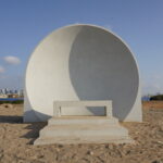 Sea Sanata: White semi-circular bench on the beach overlooking the cityscape under a clear blue sky