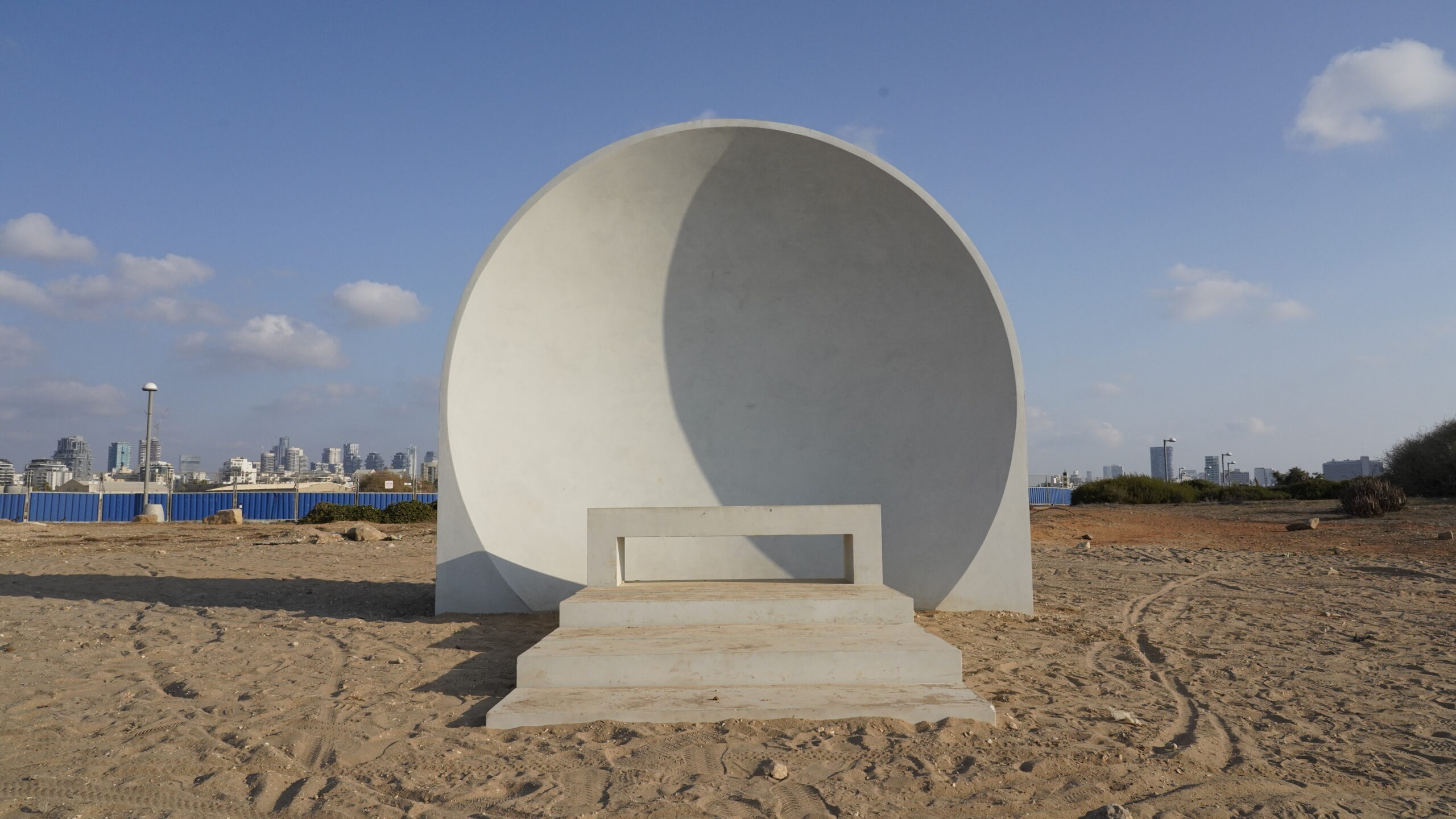 Sea Sanata: White semi-circular bench on the beach overlooking the cityscape under a clear blue sky